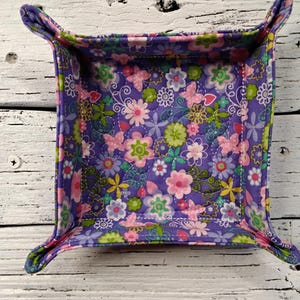 May include: A square, quilted fabric tray with a floral and butterfly pattern. The tray is primarily purple, with pink, green, and yellow flowers and butterflies. The edges are folded up to form a tray, and the corners have small, rounded tabs.