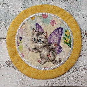 May include: A yellow fabric coaster with a white border and a whimsical design of a gray kitten with purple wings and a wand. The kitten is surrounded by flowers and a light blue sky.