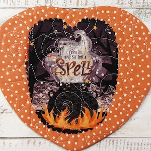 May include: Orange heart-shaped fabric pot holder with white polka dots and a stitched design. The center features a black cauldron with flames and the text "Come in and sit for a Spell".