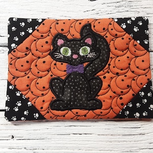 May include: A black cat with green eyes and a purple bow tie is embroidered on an orange and black quilted fabric. The fabric has a pattern of small circles and crescent moons.