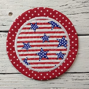 May include: A round, patriotic coaster with a red polka dot border. The center features a white and red striped pattern with blue stars. The coaster is likely made of fabric and has a stitched edge.