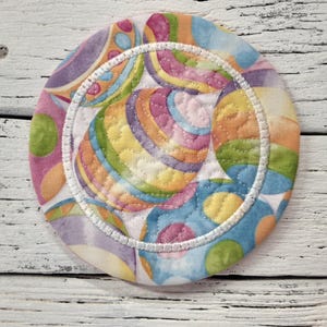 May include: A round, colorful fabric coaster with a white stitched border. The coaster features a pattern of overlapping circles in shades of pink, blue, green, yellow, and orange.