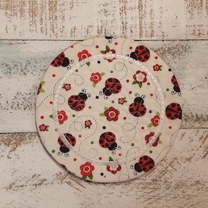 May include: A round, white fabric coaster with a quilted pattern. The coaster is decorated with red ladybugs and red flowers. The ladybugs have black spots and the flowers have green leaves. The coaster has a white stitched border.