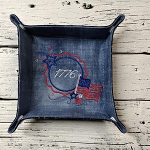 May include: A square fabric valet tray made of blue denim-like material with four corner snaps. The interior features an embroidered design with a red scalloped circle, the text 1776 in white, and a small American flag with blue and red stars.