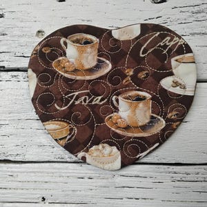 May include: Heart-shaped coffee coaster with a brown background featuring coffee cups, coffee beans, and the words "Java" and "Capp". The coaster has a quilted design with white stitching.