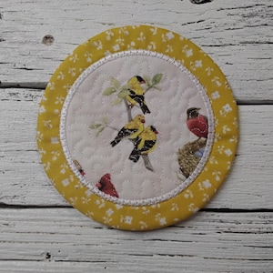 May include: A round, quilted coaster with a yellow floral border. The center features a white background with a bird and branch pattern. The coaster is approximately 4 inches in diameter.