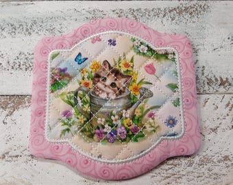 May include: A pink quilted hot pad with a white border and a floral design featuring a kitten in a watering can. The design includes flowers, butterflies, and a blue sky.