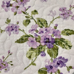 May include: A close-up of a quilted fabric with a floral pattern. The fabric is white with a light purple floral design. The flowers are small and delicate, and the fabric has a quilted texture.