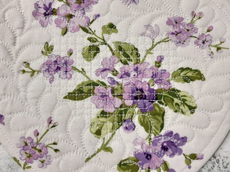 May include: A close-up of a quilted fabric with a floral pattern. The fabric is white with a light purple floral design. The flowers are small and delicate, and the fabric has a quilted texture.