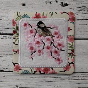 May include: Square fabric coaster with an embroidered design. The center features a bird perched on a branch of pink cherry blossoms. The border is a floral pattern with pink flowers and green leaves on a cream background.