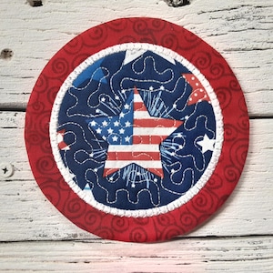 May include: A red, white, and blue fabric coaster with a star-shaped American flag design. The coaster has a white border and a red outer ring with a swirling pattern.