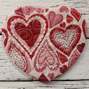 May include: A heart-shaped quilted fabric coaster with a white background and a pattern of red and pink hearts. The hearts are outlined in white stitching.