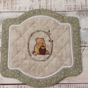 May include: A green and white quilted coaster with a brown bear embroidered in the center. The bear is sitting in a tree with a honey pot. The coaster has a scalloped edge.