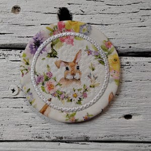 May include: A round fabric coaster featuring a bunny surrounded by a floral wreath. The coaster has a white background with a stitched border. The flowers are pink, purple, and yellow.