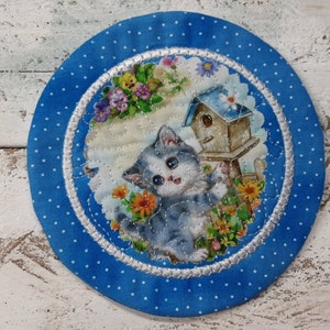 May include: A blue and white fabric coaster with a white border and a white polka dot pattern. The center of the coaster features a gray kitten with blue eyes, a birdhouse, and flowers.