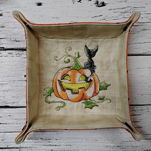 May include: A square, beige fabric tray with orange trim and corners. The tray features an embroidered Halloween design of a smiling jack-o'-lantern with a black cat on top. Green vines and leaves surround the pumpkin.
