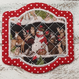 May include: A quilted coaster with a red polka dot border. The center features a winter scene with a snowman and various woodland animals. The design is stitched with white thread, creating a decorative pattern.