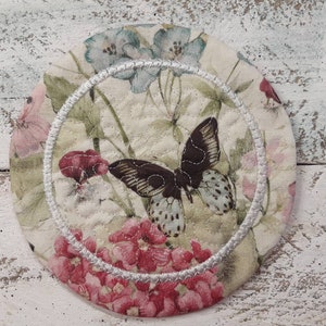 May include: Round fabric coaster with a butterfly and floral design. The coaster features a central butterfly with brown and blue wings, surrounded by pink and blue flowers on a cream background. A white stitched border frames the design.