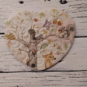 May include: Heart-shaped quilted fabric coaster with a woodland scene. The design features a tree with squirrels, birds, and a deer. The color palette includes soft pinks, greens, and browns, with a white background and decorative stitching.