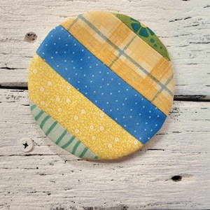 May include: A round fabric coaster with a patchwork design. The coaster features yellow, blue, and green fabric squares with different patterns.