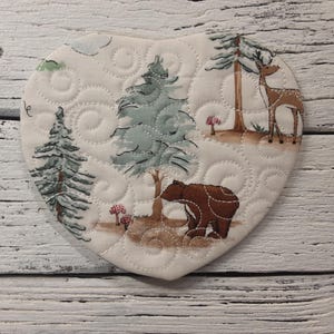 May include: A heart-shaped quilted fabric coaster with a woodland scene. The coaster features a brown bear, a deer, and pine trees on a white background with a swirling pattern.