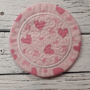 May include: Pink fabric coaster with a white border and a quilted design of hearts.