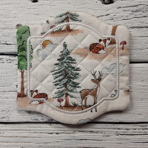 May include: A white quilted fabric coaster with a stitched border. The coaster features a woodland scene with a deer, fox, and trees.