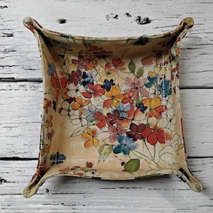 May include: A square fabric tray with a floral pattern in shades of red, orange, blue, and yellow on a cream background. The tray has four curved corners and is set against a white wooden surface.