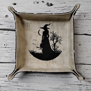 May include: A square, beige fabric tray with a black witch silhouette design. The witch is standing in front of a crescent moon and a tree. The tray has curved corners and a black border.