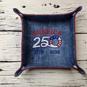 May include: A square, denim-like tray with red trim and four corner tabs. The tray features embroidered text that reads "AMERICA 250 1776-2026" in red and white, with a circular design incorporating the American flag.