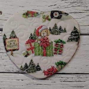 May include: A heart-shaped quilted fabric coaster featuring a printed scene of a dog wearing a Santa hat surrounded by wrapped gifts and evergreen trees. A sign on the left reads Happy Holidays. The fabric has white swirl stitching throughout.