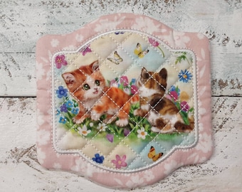 May include: A pink quilted hot pad with a white embroidered border features a vintage illustration of two kittens in a field of flowers and butterflies.