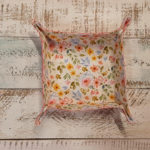 May include: A square fabric tray with a floral pattern of pink, yellow, and blue flowers on a white background. The tray has a pink border and four corner tabs. The tray is on a white wooden surface.