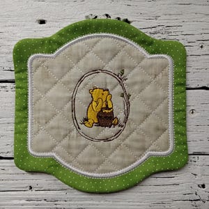 May include: Embroidered Winnie the Pooh pot holder with a green polka dot border. The center features a yellow Winnie the Pooh character with a honey pot inside a brown oval frame. The pot holder has a quilted design.