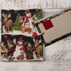 May include: A black fabric wallet with a winter woodland scene featuring snowmen, deer, and owls. The wallet has a red button closure and a beige fabric interior with a red fabric pocket.