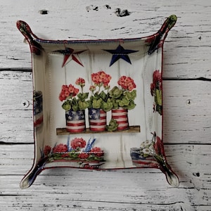May include: Square decorative tray with a patriotic theme. The tray features a design of red, white, and blue elements, including stars, stripes, and floral arrangements in pots. The tray has a white background and a red border.