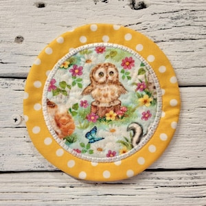 May include: Yellow fabric coaster with white polka dots and a white border. The center features a colorful print of an owl, butterfly, and other woodland creatures.