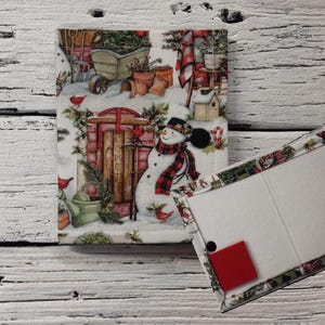 May include: A white fabric notebook cover with a winter scene featuring a snowman, red cardinals, and a red and black plaid scarf. The cover has a white fabric insert with a red fabric accent.