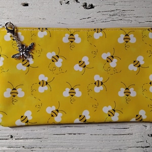 May include: Yellow zippered pouch with a repeating pattern of cartoon bees and white clouds. A silver bee charm hangs from the zipper pull. The pouch is rectangular and has a white zipper.