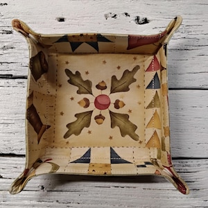 May include: A square, decorative tray with a painted, quilted design. The tray features a central floral motif with acorns and leaves, surrounded by patchwork patterns in shades of beige, brown, blue, and red. The tray has four curved handles.