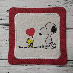May include: Square quilted coaster featuring Snoopy and Woodstock with a red heart balloon. The coaster has a red border with a swirl pattern and a white quilted center. A charming, decorative item.