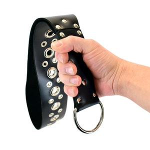May include: A black leather belt with silver metal eyelets and a silver metal ring. The belt is held in a hand.