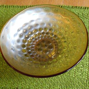 Pu&ograve; includere: Una ciotola decorativa in vetro con un interno strutturato e increspato. La ciotola ha una tonalit&agrave; dorata con un bordo marrone scuro. La ciotola &egrave; su un sottobicchiere verde intrecciato.