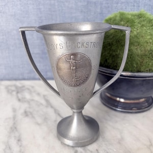 May include: A silver-toned metal trophy cup with two handles, featuring an engraved circular medallion of a swimmer. The text reads BOYS BACKSTROKE, 1938, THE BEACH CLUB, and 1ST PRIZE. The trophy has a smooth, tapered body and a circular base.