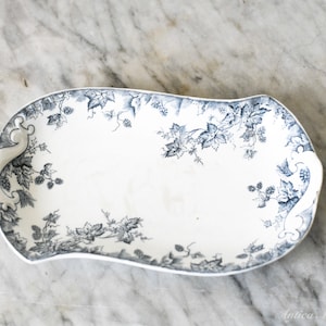 May include: A white ceramic serving platter with a blue floral design. The platter has a unique, curved shape and is decorated with a detailed pattern of leaves and vines around the edges. The platter is approximately 30 cm long.