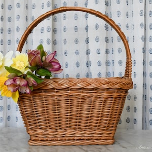 May include: A woven brown wicker basket with a handle, filled with yellow, white, and purple flowers. The basket has a rectangular base and a braided rim. The handle arches over the top. The background features a white curtain with a blue floral pattern.