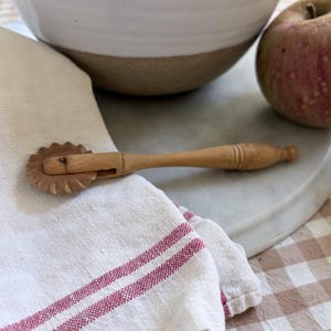 Könnte beinhalten: Ein Teigrad aus Holz mit einem gezackten, runden Schneider und einem schlanken, gedrechselten Griff. Das Werkzeug hat eine natürliche, hellbraune Holzmaserung, einen kleinen Metallstift zur Befestigung und ein schlichtes Design.