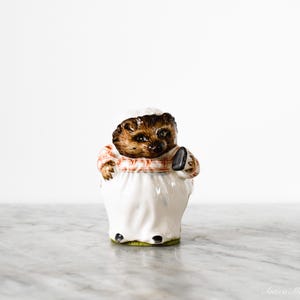 May include: A ceramic figurine of a hedgehog dressed as a chef. The hedgehog has brown fur, a white chef's hat, and a red and white checkered scarf. It wears a white apron and holds a black utensil.
