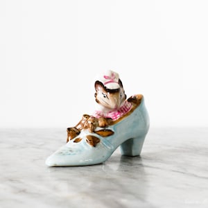 May include: A vintage porcelain figurine featuring a mouse wearing a pink and white outfit, seated in a light blue high-heeled shoe. The shoe has brown accents and a pointed toe. The figurine is set against a white background.