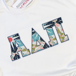 May include: A white sweatshirt with the letters "AΛI" embroidered on the front. The letters are filled with a colorful floral pattern and outlined in navy blue. The brand name "monogram A-LA-MODE" is printed on the bottom right.